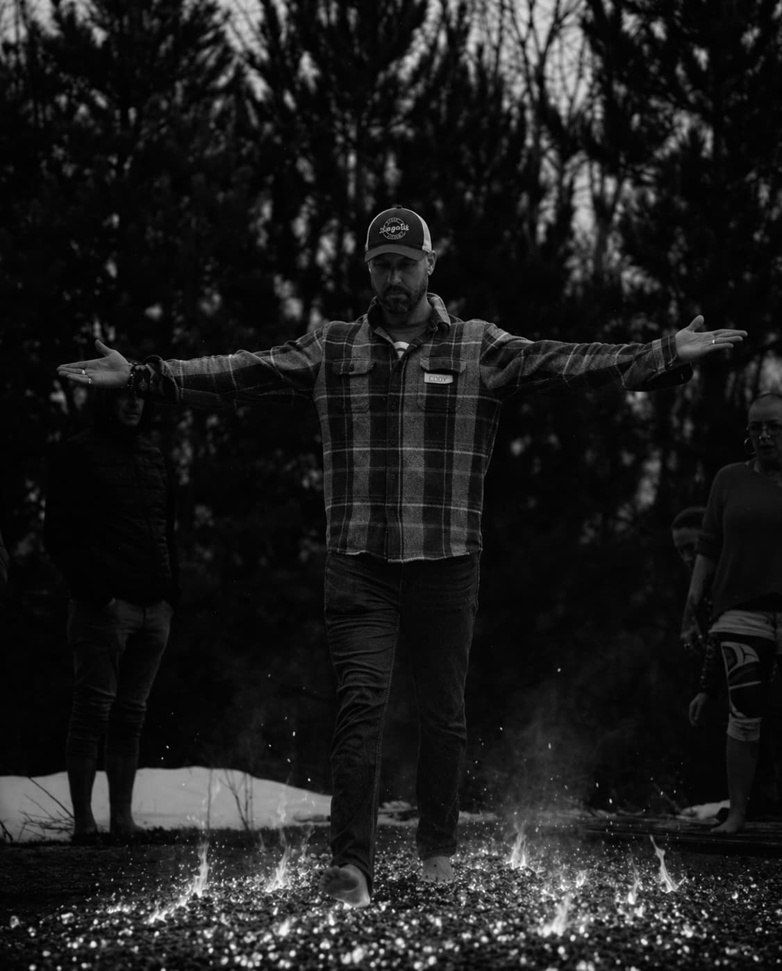 Man in a plaid shirt walks barefoot across glowing hot coals with arms outstretched.