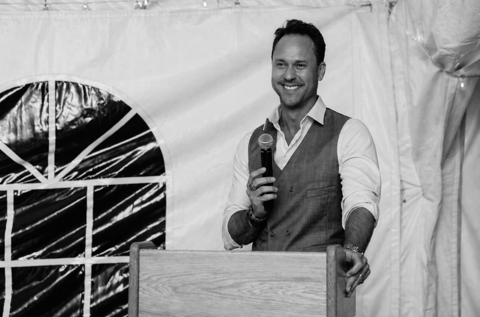 Smiling man in a vest holding a microphone at a wooden podium inside a tent.