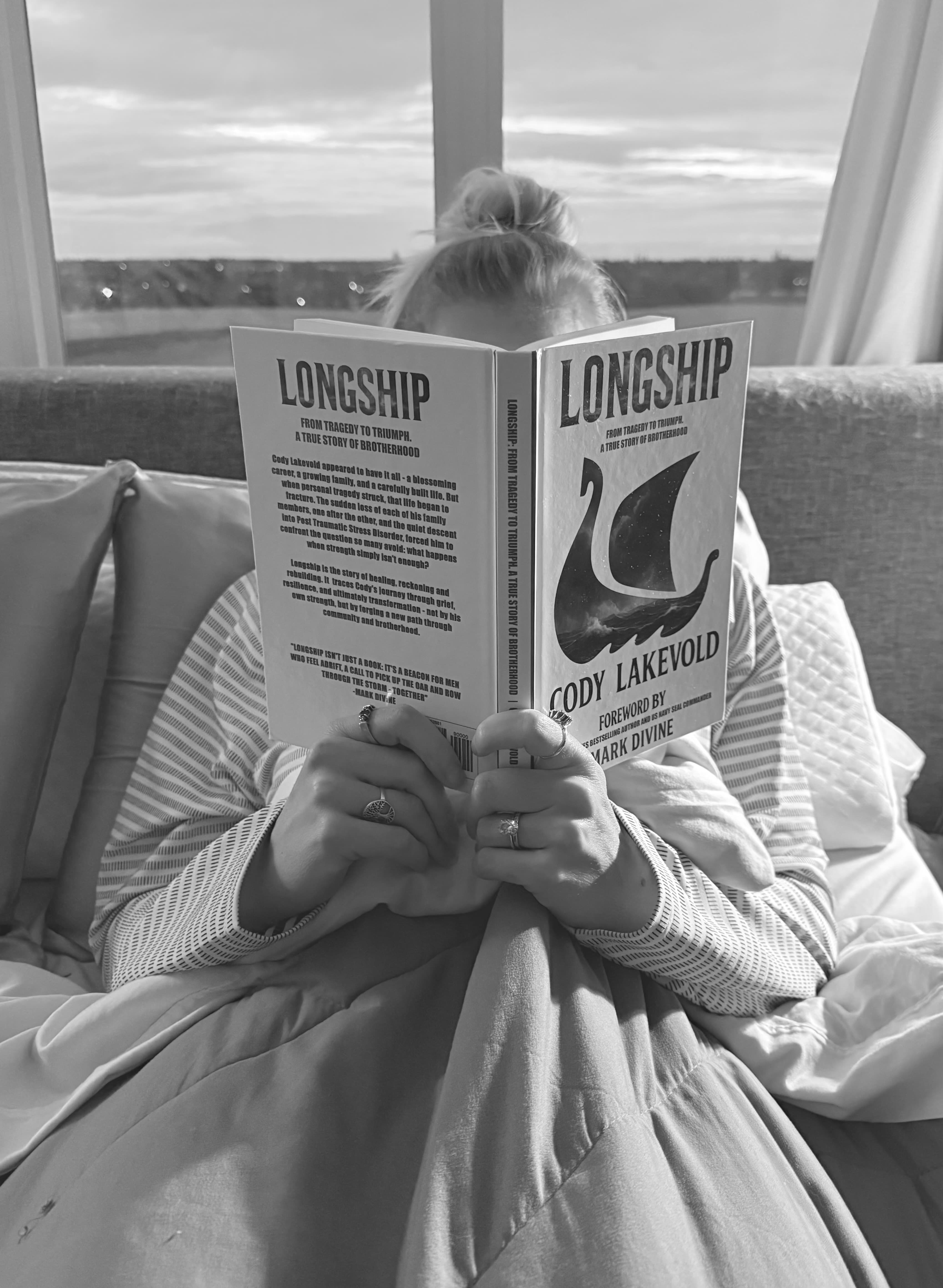 A person in bed holds the book Longship by Cody Lakevold, obscuring their face.