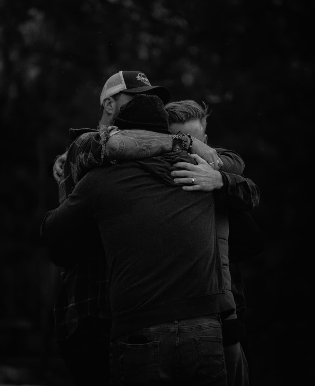 Three men sharing a tight, supportive group hug in a black and white setting.