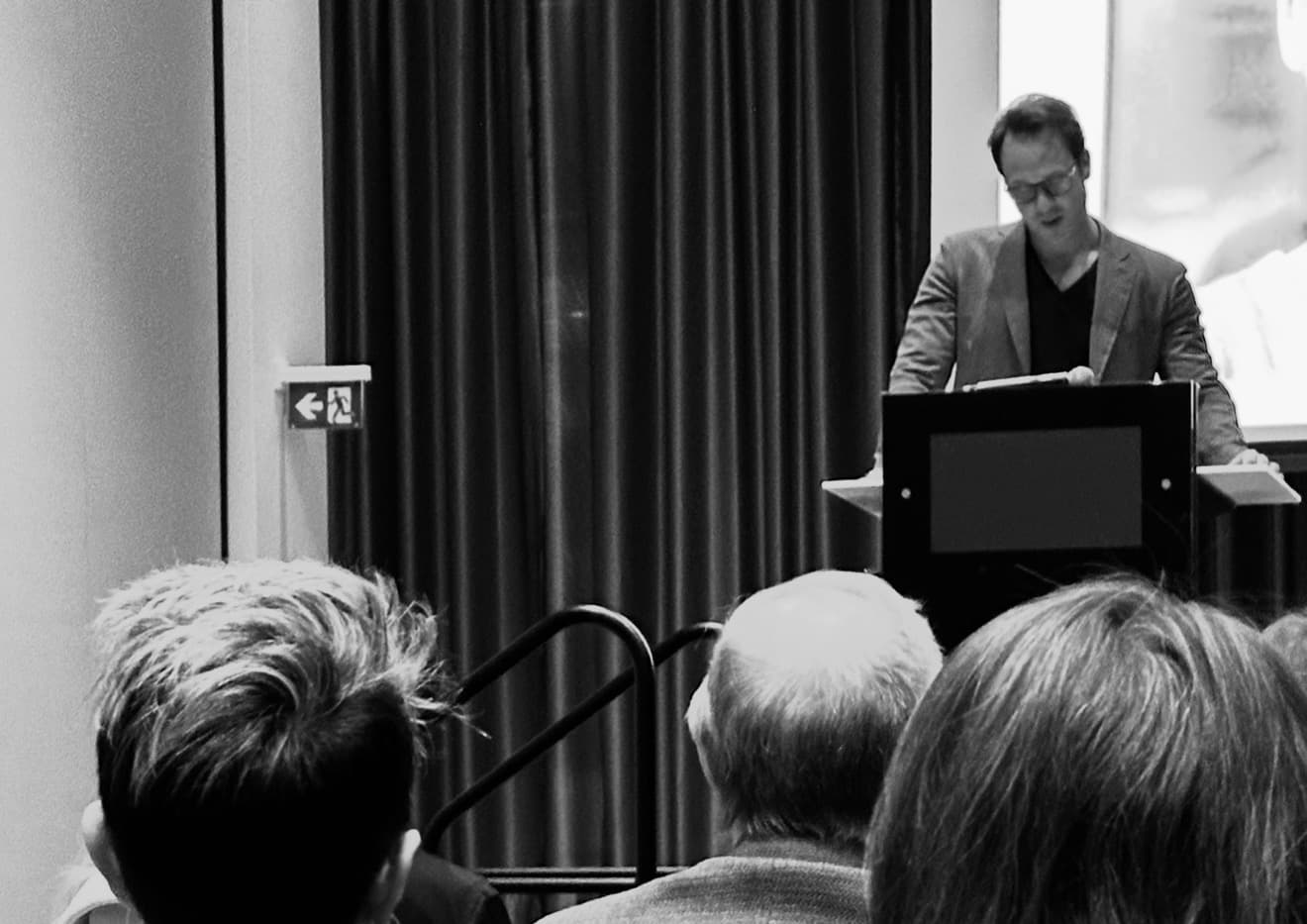 Man in a blazer speaks at a podium before a seated audience in monochrome.