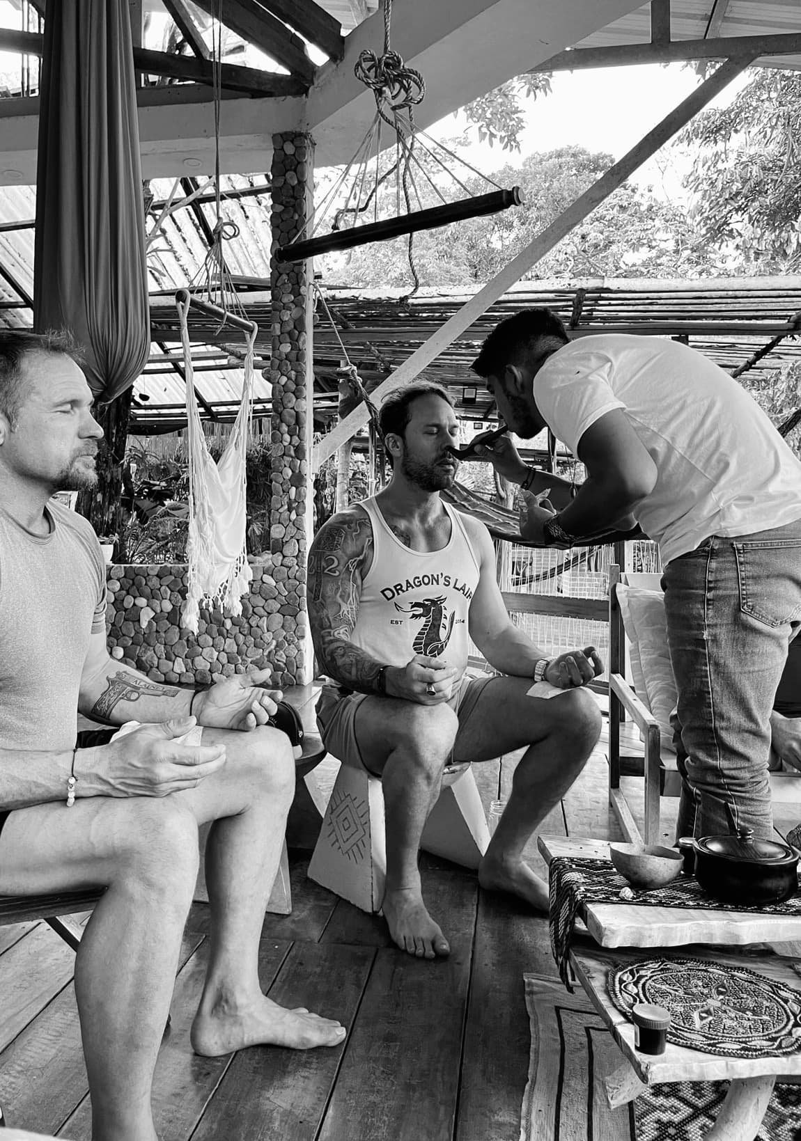 Man administering traditional nasal medicine to a seated man in a rustic outdoor pavilion.